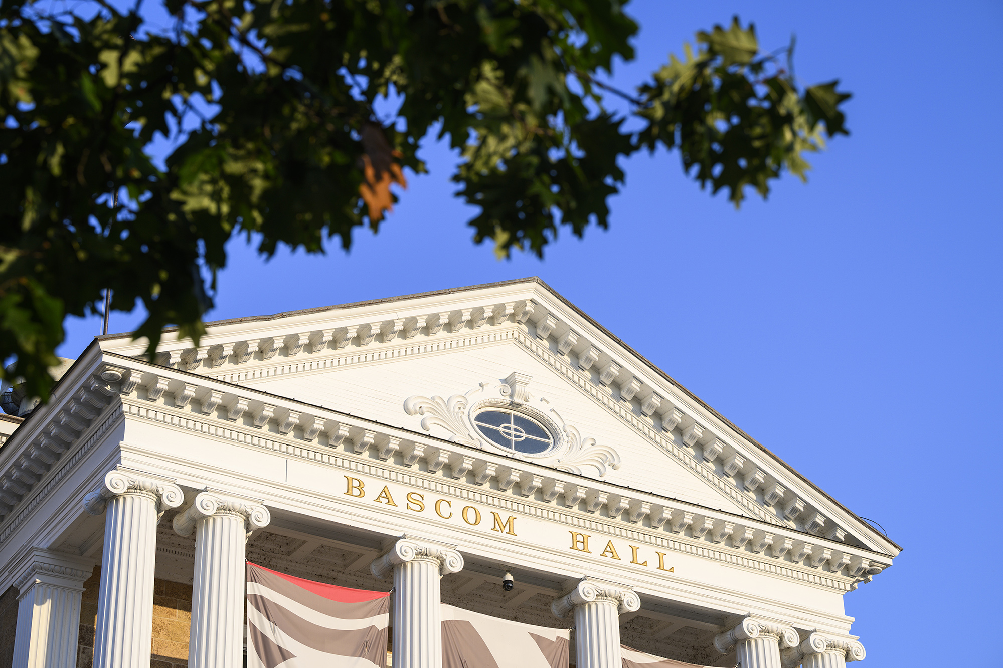 The front of Bascom Hall with white columns and a gold nameplate, framed by green leaves against a clear sky.