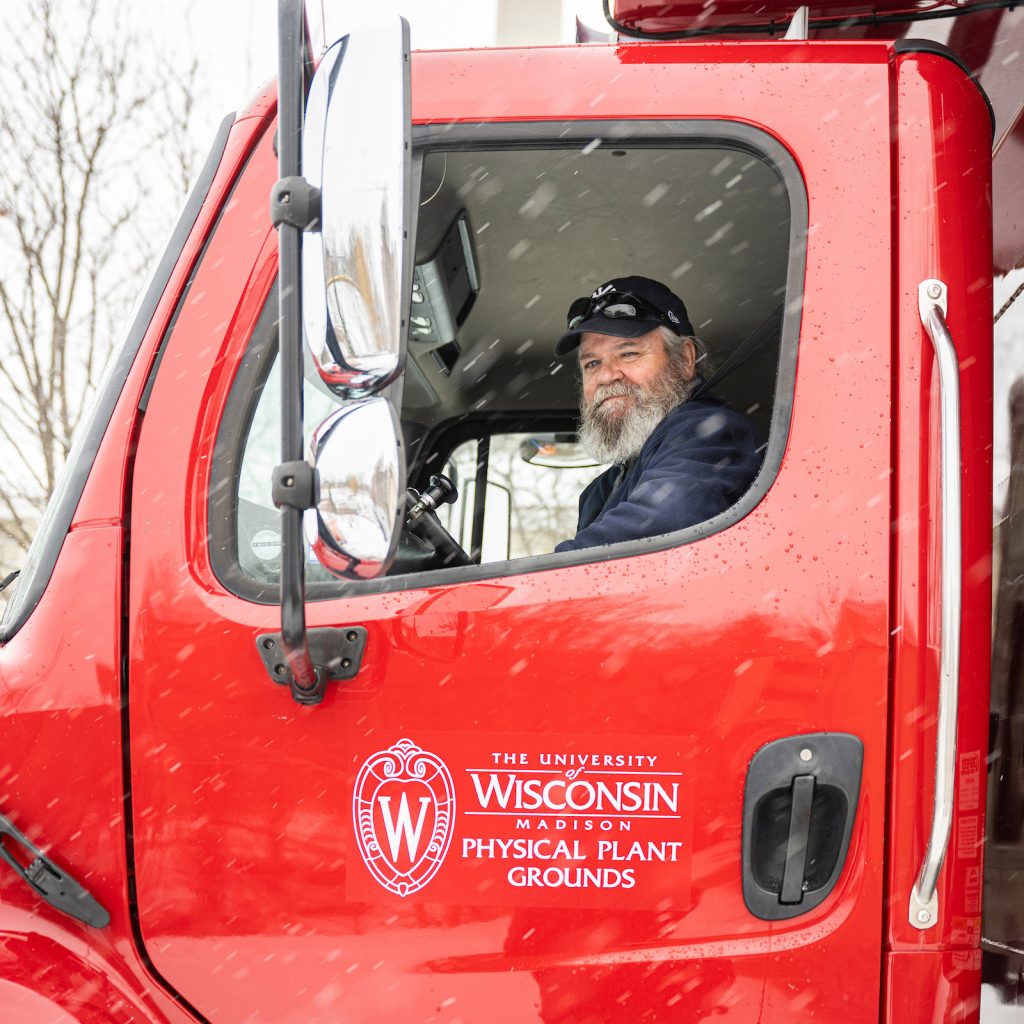 A person sitting in the cab of a red truck. The side of the truck has a UW–Madison logo with the text "Physical Plant Grounds."