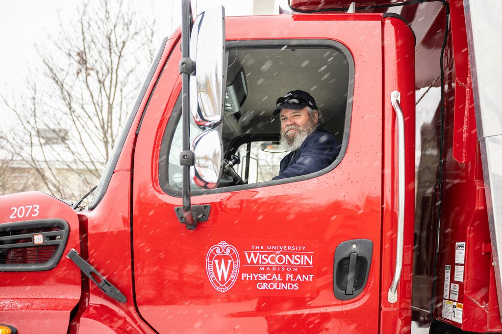 A person sitting in the cab of a red truck. The side of the truck has a UW–Madison logo with the text "Physical Plant Grounds."