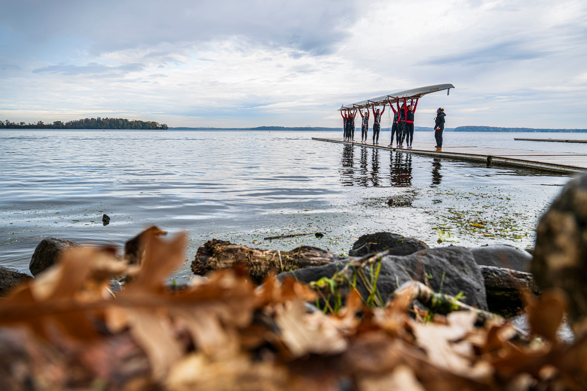 Rowers on a dock lifting a long racing boat overhead, with calm water and a distant tree‑covered shoreline behind them.